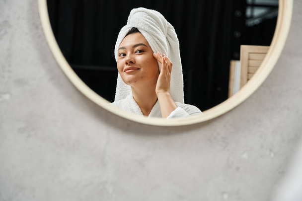 молода азіатка з білим рушником на голові торкається шкіри акне і дивиться на дзеркало у ванній кімнаті - Фото, зображення