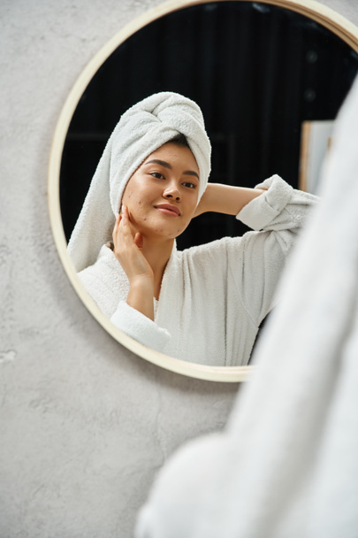 mujer asiática joven con toalla blanca en la cabeza y piel propensa al acné mirando el espejo del baño en casa - Foto, Imagen