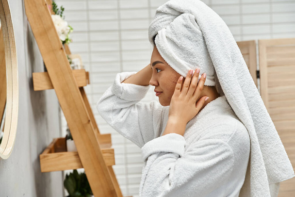 Seitenansicht der jungen asiatischen Frau mit Handtuch auf dem Kopf und Akne neigender Haut beim Blick auf den Badezimmerspiegel - Foto, Bild