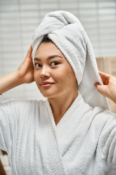 pretty asian woman in robe with white towel on head looking at camera in bathroom, skin issues - Фото, изображение