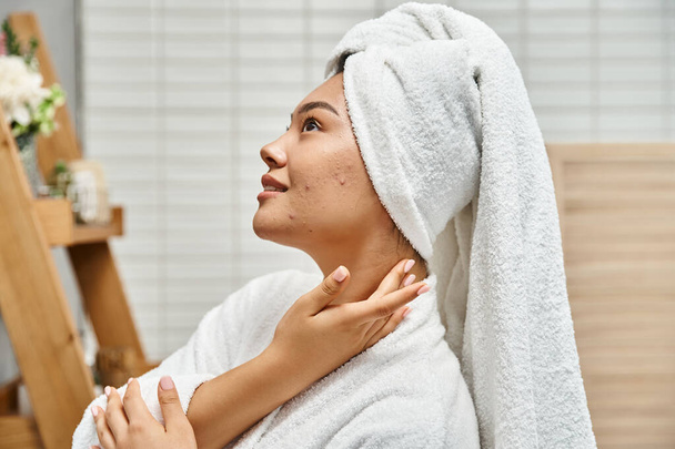 pleased young asian woman with acne prone skin with white towel on head posing in bathroom - Photo, Image
