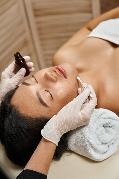 huidverzorging, therapeut die behandelingsserum met wattenstaafje toepast op Aziatische vrouw met een acne-gevoelige huid - Foto, afbeelding
