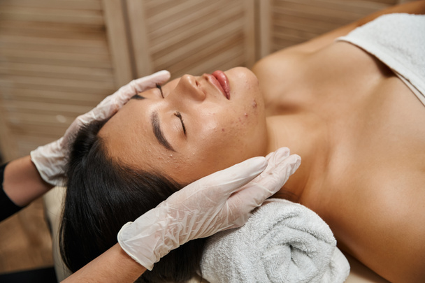 masaż twarzy dla skóry trądzikowej, terapeuta w rękawiczkach lateksowych i azjatycki klient z zamkniętymi oczami - Zdjęcie, obraz
