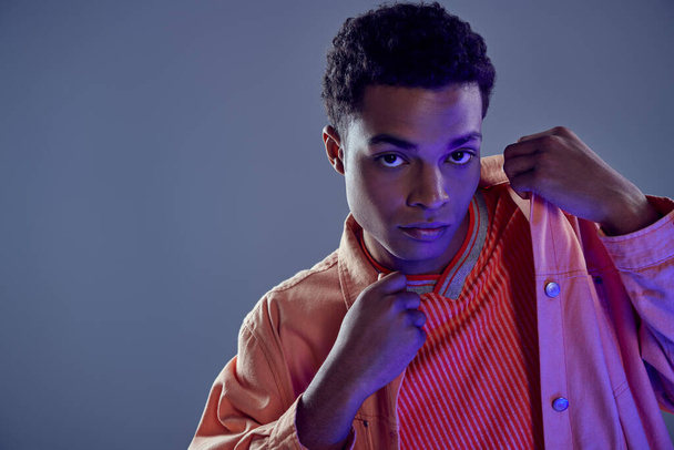 retrato de chico afroamericano guapo en camisa de melocotón mirando a la cámara en gris con luz azul - Foto, imagen