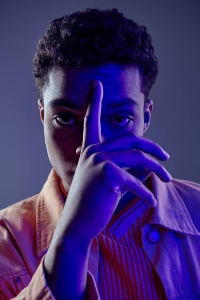 Şeftali rengi gömlekli Afro-Amerikan bir adam. Yüzüne yakın parmaklarıyla gri ışıkta kameraya bakıyor. - Fotoğraf, Görsel