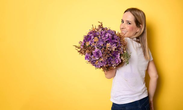Retrato de una hermosa joven vestida con ropa casual sosteniendo un gran ramo de iris y tulipanes aislados sobre fondo amarillo - Foto, imagen