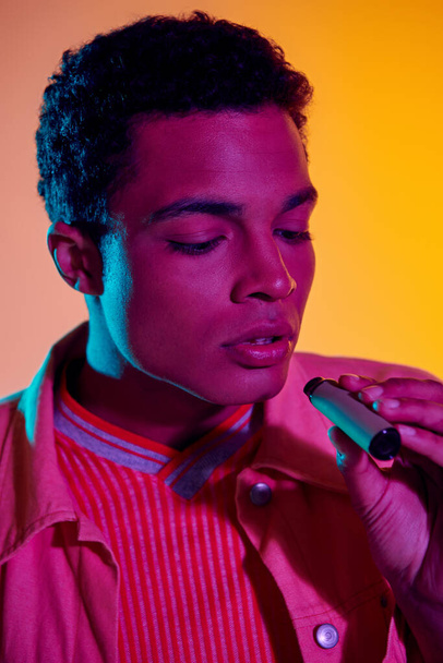 african american man vaping with electronic cigarette in hand, orange backdrop with lighting - Photo, Image