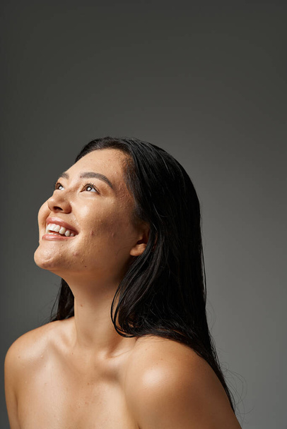 junge asiatische Frau mit brünetten Haaren und zu Akne neigender Haut beim Anblick des Spiegels im Badezimmer, Banner - Foto, Bild