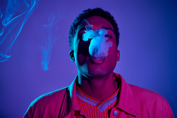 stylischer afrikanisch-amerikanischer Kerl, der Rauch vor blauem Hintergrund mit violettem Licht ausatmet, gen z - Foto, Bild