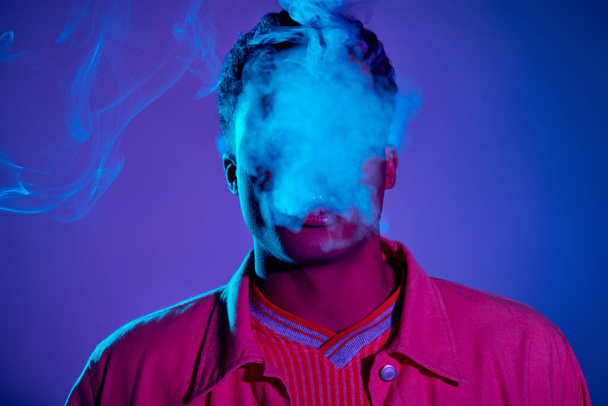 retrato del hombre afroamericano exhalando humo sobre fondo azul con iluminación púrpura, gen z - Foto, imagen