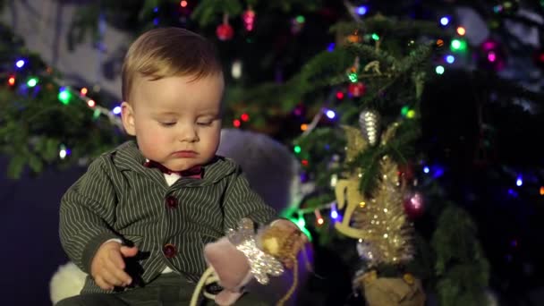 Het kind zit bij de kerstboom. Feestelijke sfeer, nieuwjaars- en kerstboom. Het jongetje is mooi gekleed en heeft blond haar. Kerstmis voor kinderen, gelukkig nieuwjaar - Video