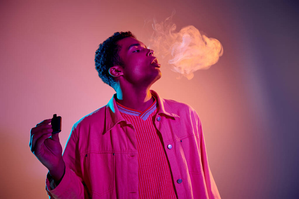 portrait of african american man exhaling smoke while holding e cigarette on colorful background - Photo, Image