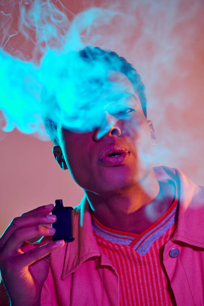 portrait of african american guy exhaling smoke while holding e cigarette on vibrant background - Photo, Image