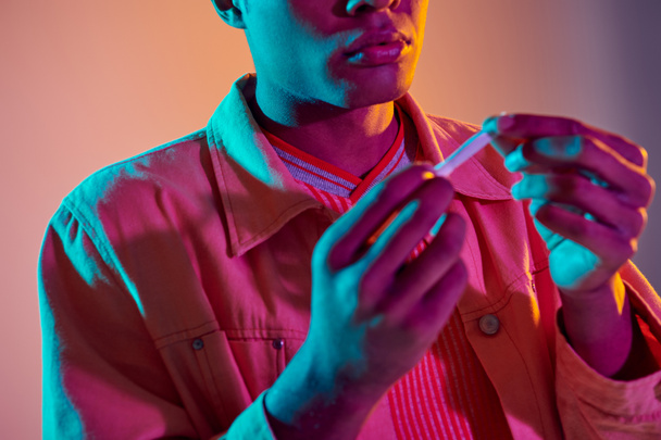 обрізаний афроамериканський хлопець тримає рулонну сигарету на барвистому фоні з блакитним неоновим освітленням - Фото, зображення