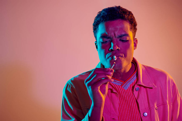 focalizzato ragazzo africano americano illuminazione sigaretta con partita sotto luci al neon blu su sfondo rosa - Foto, immagini