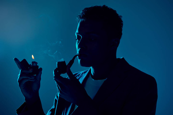 giovane uomo afro-americano che tiene più leggero e fumante pipa su sfondo blu scuro con illuminazione - Foto, immagini