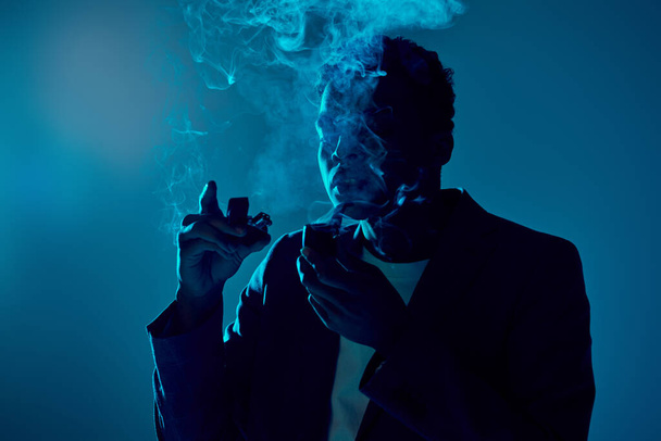 krullend afrikaanse amerikaanse man met aansteker en pijp, terwijl het uitademen van rook op donkerblauwe achtergrond - Foto, afbeelding