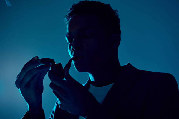curly african american man holding lighter while smoking pipe on dark blue backdrop with lighting - Photo, Image