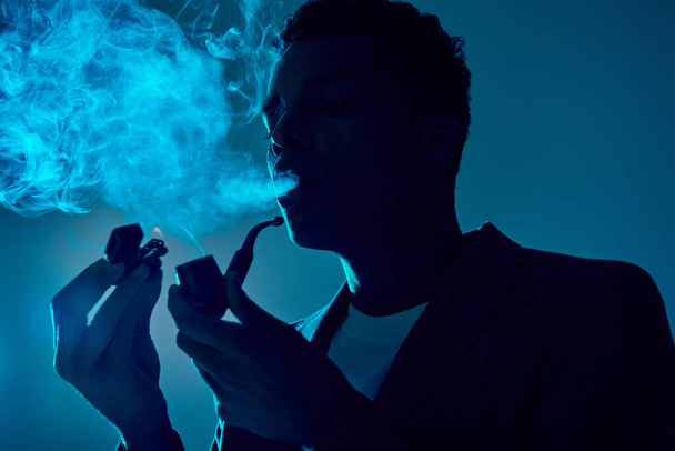 afro americano cara segurando isqueiro e tubo enquanto exalando fumaça no fundo azul escuro - Foto, Imagem