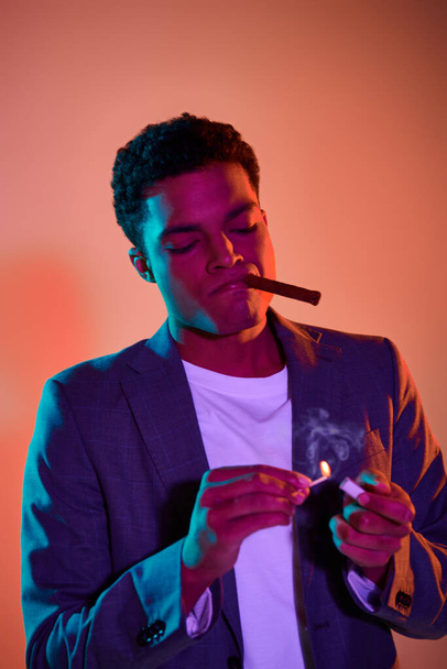 african american guy with cigar holding match with fire on pink background with blue lighting - Photo, Image