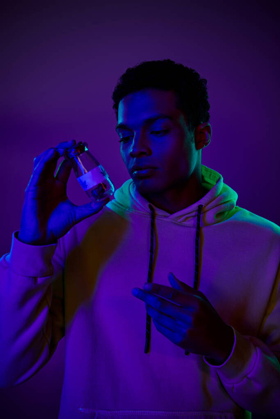 jonge Afro-Amerikaanse man in hoodie met pot met medicinale cannabis op donkerblauwe achtergrond - Foto, afbeelding