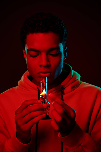 young african american man in hoodie lighting glass bong on dark background with red lighting - Photo, Image