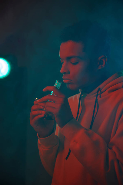 humo alrededor afroamericano chico en sudadera con capucha celebración de vidrio bong sobre fondo oscuro, cannabis medicinal - Foto, imagen