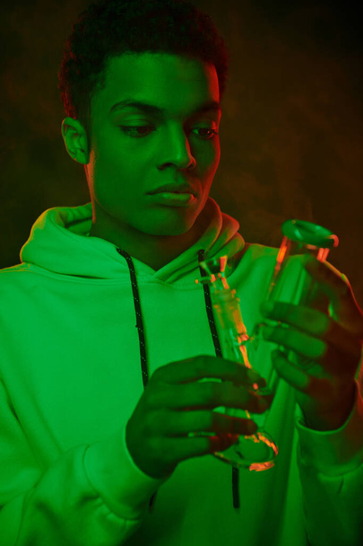 young african american man in hoodie looking at glass bong on dark background with green lighting - Photo, Image