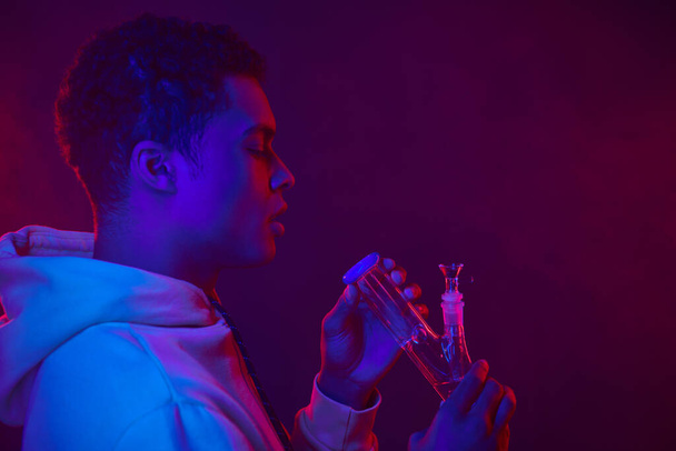 junger afrikanisch-amerikanischer Mann in Kapuzenpullover mit Blick auf Glasbong auf dunkelviolettem Hintergrund mit Beleuchtung - Foto, Bild