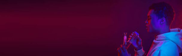 молодой африканский американец в толстовке смотрит на стеклянный бонг на темно-фиолетовом фоне, баннер - Фото, изображение