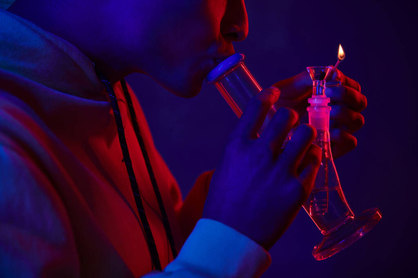 Kırpılmış Afro-Amerikan kapüşonlu adam koyu mavi arka planda kırmızı ışıklı cam bong yakıyor. - Fotoğraf, Görsel