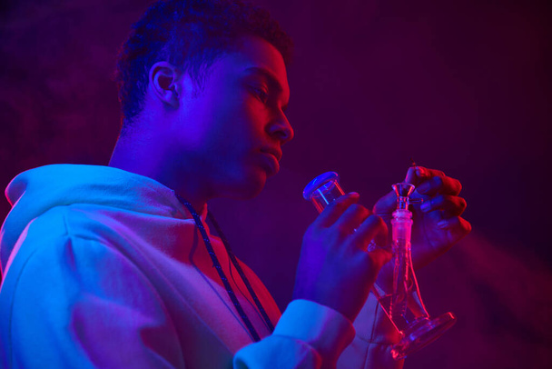 молодий афроамериканський чоловік в светрі освітлює скляний бонг на темно-синьому фоні з червоним світлом - Фото, зображення