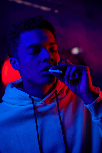Elinde sigara tutan Afro-Amerikan bir adam koyu mavi arka planda kırmızı ışıkla ağzının yanında sarkmış sigara tutuyor. - Fotoğraf, Görsel