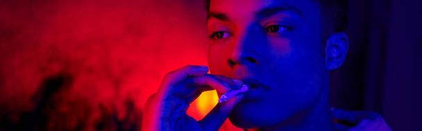 african american man holding rolled cigarette near mouth on dark blue with red light, smoker banner - Photo, Image