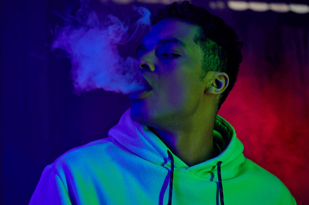 poważny african amerykański człowiek wydychając dym na ciemnym tle z niebieskim i czerwonym światłem, palacz - Zdjęcie, obraz