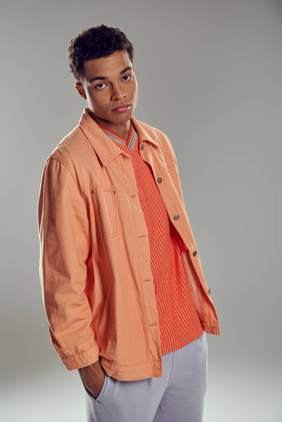 young african american man in peach color shirt looking at camera on grey backdrop, hand in pocket - Foto, afbeelding