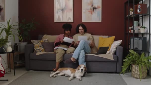 Longo tiro de jovem casal interracial olhando através de contas enquanto contando seu orçamento juntos, sentado no sofá com laptop e cão beagle na sala de estar moderna - Filmagem, Vídeo