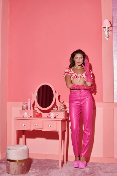 鏡付きの虚栄心のテーブルの近くに立っている間に人形のようにポーズするピンクの服の美しい若い女性 - 写真・画像