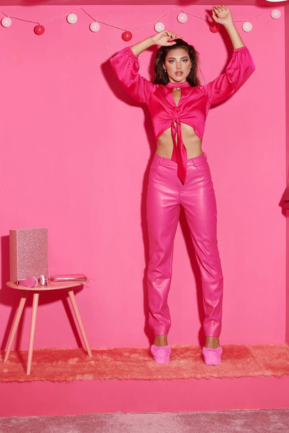 mujer joven glamorosa en top de la cosecha y pantalones de color rosa de pie con las manos levantadas cerca de la guirnalda bola - Foto, Imagen