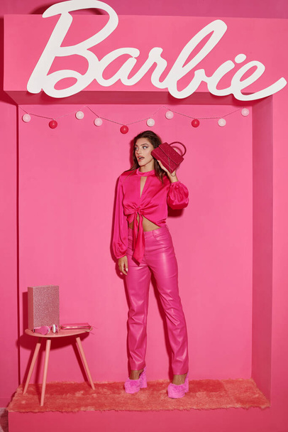 KYIV, UKRAINE - HEINÄKUU 14, 2023: hämmästynyt ja nukke kuin nainen sato alkuun ja vaaleanpunainen housut seisoo tyylikäs käsilaukku lähellä Barbie merkki - Valokuva, kuva