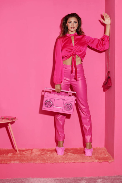 gyönyörű fiatal nő élénk öltözékben gazdaság retro boombox és integető kéz rózsaszín háttér - Fotó, kép