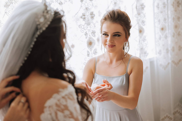 Morning of the bride. The bride's maid of honor helps the bride lace up her dress, fasten buttons on the dress or sleeves. Girlfriends help the bride fasten her dress - Fotó, kép