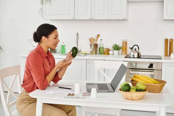 felice nutrizionista afroamericano che tiene avocado maturo e dà consigli dietetici sul laptop in cucina - Foto, immagini