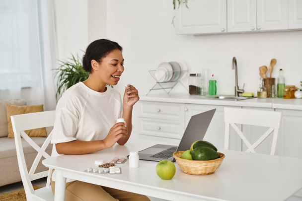 Mutlu Afro-Amerikan beslenme uzmanı laptopun önünde ilaç ve meyve alıyor. - Fotoğraf, Görsel