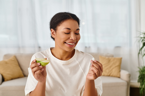 fröhliche afrikanisch-amerikanische Ernährungswissenschaftlerin vergleicht Nahrungsergänzungsmittel mit frischem Apfel für eine gesunde Ernährung - Foto, Bild
