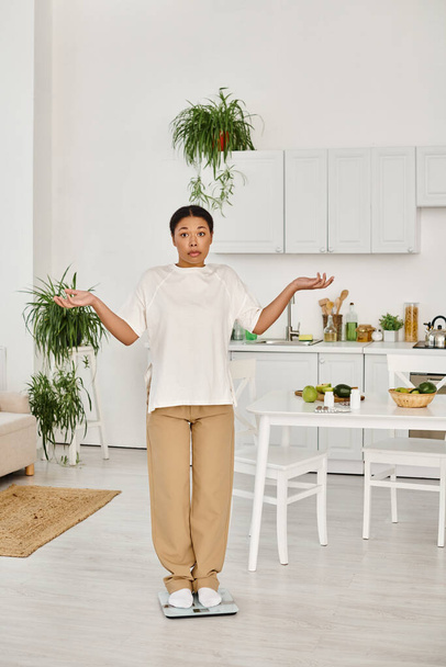 confused black woman in casual attire standing on scale in kitchen, weight management routine - Photo, Image