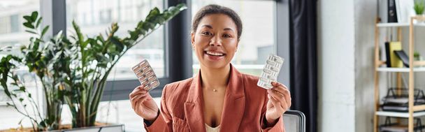 nutricionista afroamericano alegre mostrando diferentes medicamentos mientras está sentado en la oficina, pancarta - Foto, imagen