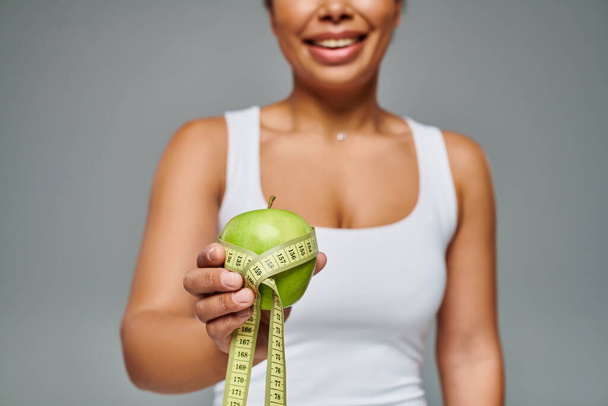 cropped view of happy african american woman with measuring tape and apple on grey background - Photo, Image