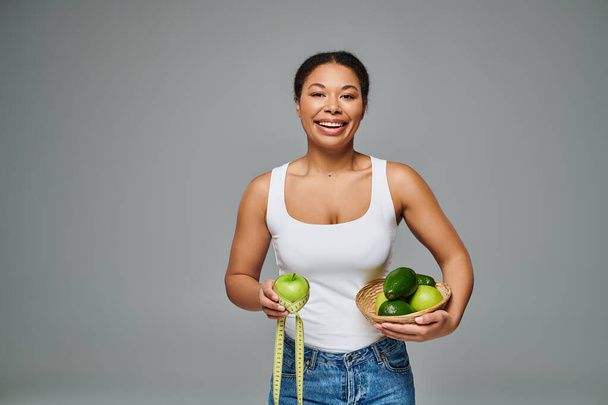 heureuse diététiste afro-américaine avec pomme et suppléments mettant en valeur la santé sur fond gris - Photo, image