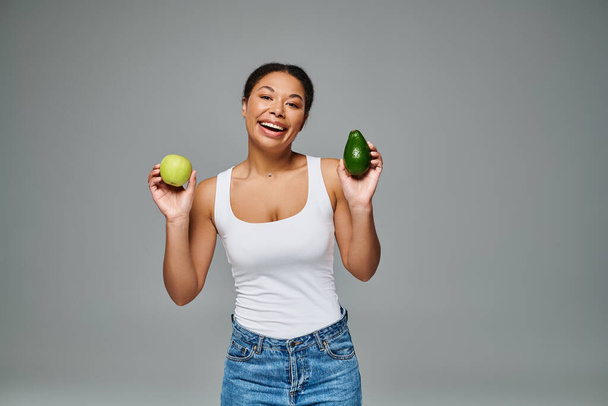 Сияющая африканская американка с яблоком и авокадо, способствующая сбалансированному питанию на сером фоне - Фото, изображение